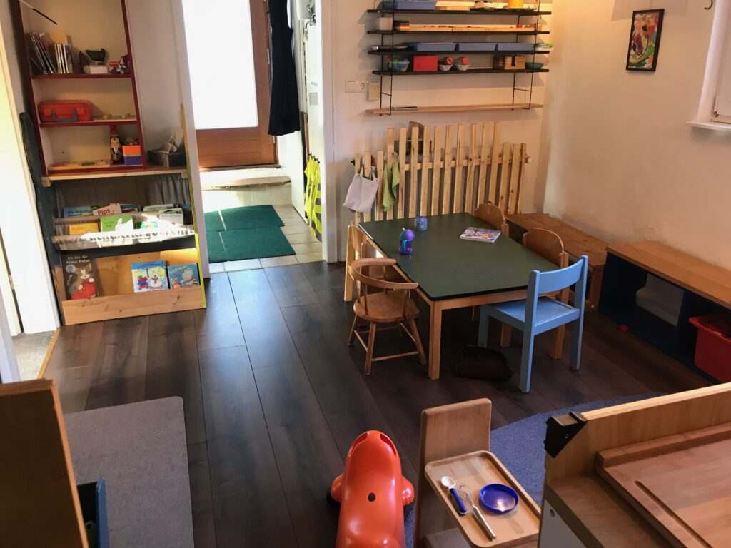 Einblick in einen der Räume der Kindertagespflege. Zu sehen ist rechtsseitig ein grünbeschichteter Tisch mit fünf Kinderstühlen. Dahinter an der Wand ist ein Regal mit verschiedenen Arbeitstabletts an der Wand. Linksseitig ist ein Regal mit zwei Ebenen in Griffhöhe der Kinder gefüllt mit Büchern. Weiter oben im Regal stehen weitere Spielmaterialien und oben Infomaterial für die Eltern. Mittig ist der Durchgang zum Flur, die Haustür und ein Teil der Garderobe zu sehen.