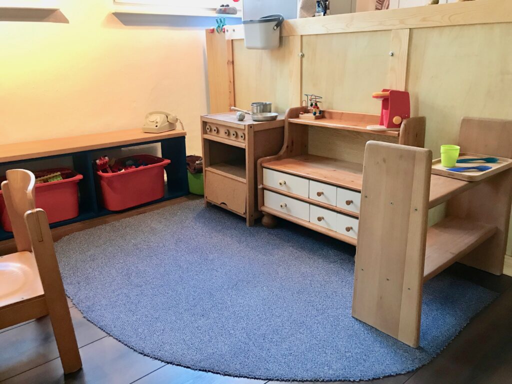 Auf der linken Seite ist ein niedriges stuhlhohes Regal zu sehen in der zwei rote Boxen mit Duplo-Bausteinen stehen. Oben auf dem Regal steht ein Wählscheiben-Telefon. Im rechten Winkel dazu zur Bildmitte hin ist ein Spielherd aus Holz mit kleiner Pfanne und kleinem Topf aus Metall darauf zu sehen. Daneben schließt sich eine niedrige Küchenkommode aus Holz an. Sie hat vier kleine und zwei große Schubladen in weiß und ein höheres Bord auf dem eine Spiel-Kaffeemaschine steht. Am rechten Bildrand ist ein zweietagige kleines Regal erkennbar. Die Möbel sind so gestellt, dass sie eine Art "U" bilden. In diesem "U" liegt ein blauer Teppich. Den Hintergrund der Küchen- und Bauecke bildet die Holztrennwand zur "echten" Küche.