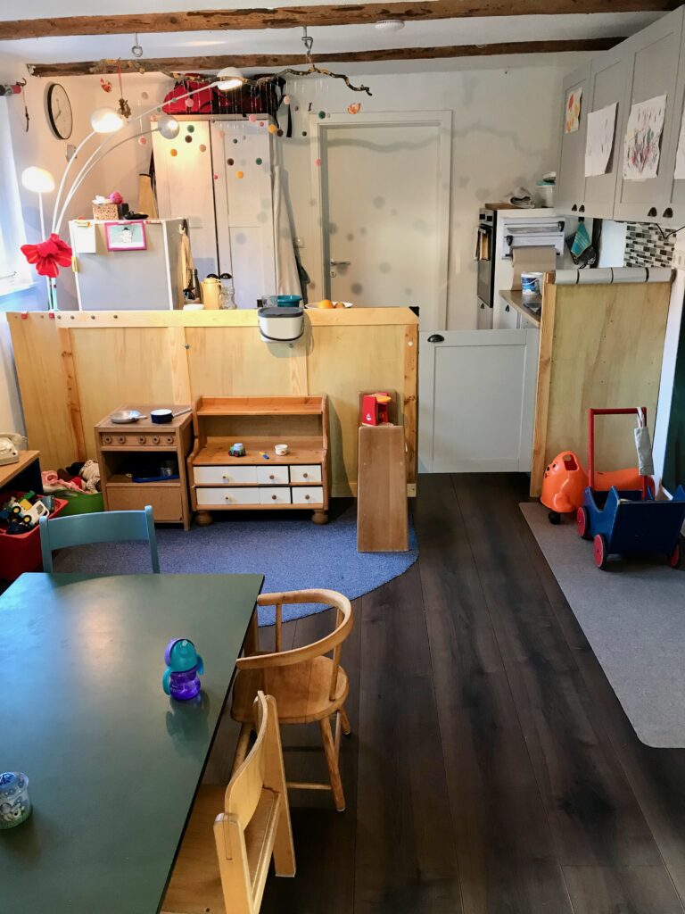 Hier ist eine Totale auf den Ess-, Bau- und Bastelbereich zu sehen. Im Vordergrund befindet sich der Kindertisch (vgl. "Essecke"), linksseitig die Bau- und Küchenecke für die Kinder (vgl. "Kinderküche/ Bauecke"), rechtseitig sind ein Puppenwagen aus Holz und eine Rutschrobbe zu sehen. Im Hintergrund des Bildes lassen sich eine Küchenarbeitplatte ein Herd und Küchenschränke erahnen. Der Küchenbereich ist durch eine graue niedrige Tür gesichert.