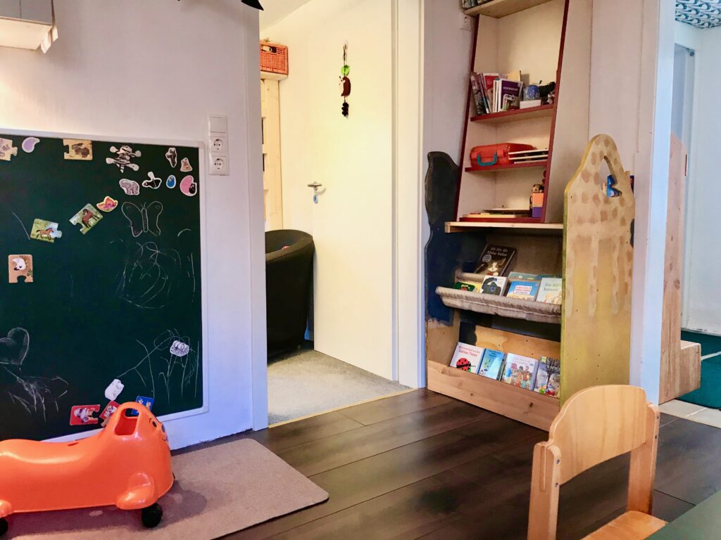 Auf diesem Bild ist mittig das Leseregal mit seinen Hängematten-Fächern aus beigem Stoff, dem bodennahen Stehfach und dem Aufbau mit weiterem Material für Kinder und Eltern (im oberen Bereich) erfasst. Erkennbar ist der Durchgang zum Garderobenbereich rechtseitig und links vom Bücherregal die Tür zum Toberaum. Ganz links im Bild ist die große Wandtafel, auf der zahlreiche Magneten kleben. Vor der Tafel ist ein hellbrauner Teppich erkennbar und eine Rutschrobbe. Im Vordergrund ist der Esstisch der Kinder und ein Stuhl erahnbar.