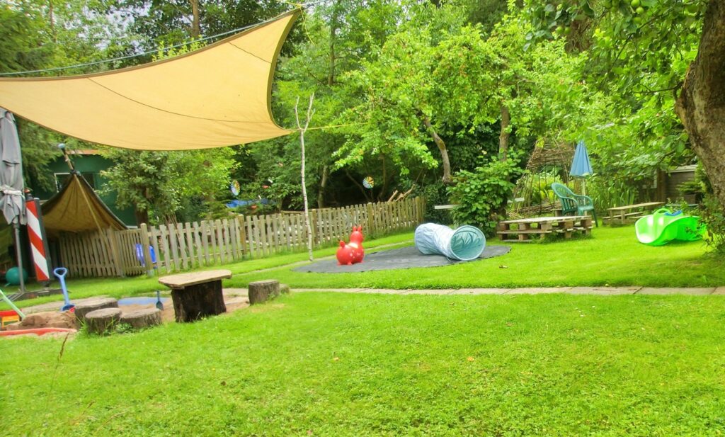 Dieses Bild zeigt eine große Rasenfläche, eingerahmt durch Bäume und Büsche. Linker Hand ist eine Sandkiste mit Holztisch und Stammabschnitten als Sitz- und Klettermöglichkeit zu erkennen. Über der Sandkiste hängt ein hellbraunes Sonnensegel. In der Bildmitte liegt ein rundes Trampolinsprungtuch. Auf dem Sprungtuch (die Manege) steht ein rotes Rowdy neben einem hellblauen Kriechtunnel. Rechts ist noch eine grüne Wippschaukel aus Plastik zu erahnen.
