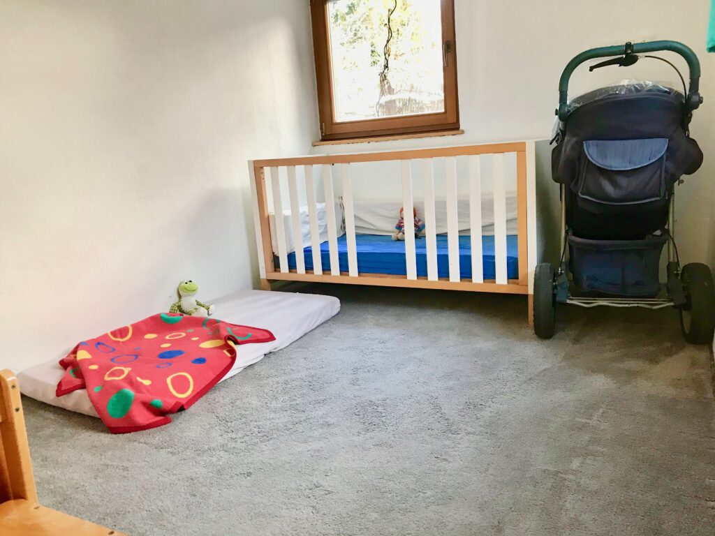 Dieses Bild zeigt den kleinen Schlafraum von der Tür aus. Es steht eine blaue Karre rechtsseitig neben einem Kinderbett. Das Kinderbett hat ein Durchschlupfloch für die Kinder zu selbst ein- und aussteigen. Über dem Bett ist ein Fenster in den Garten. Links vor dem Bett, im "L" angeordnet, liegt noch eine Kindermatratze auf der eine rot-bunte Decke liegt.