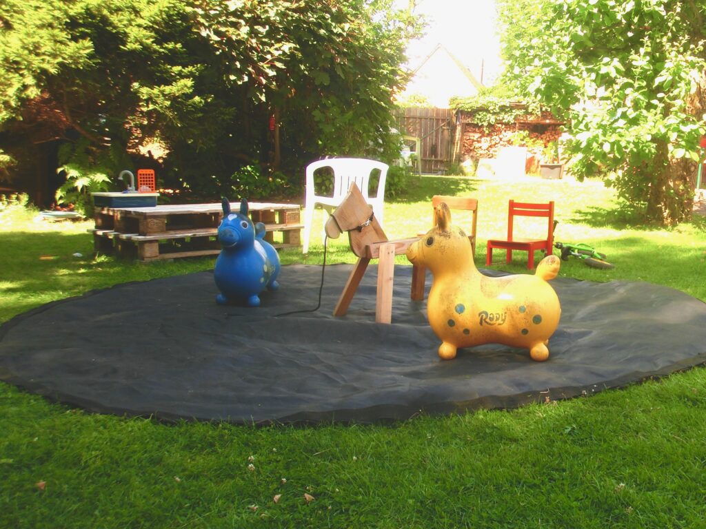 Luftig eingerahmt von Büschen liegt auf einer Rasenfläche das Sprungtuch eines Trampolins ausgereitet (die Manege). Im Hintergrund am Rand des Tuchs stehen Kinderstühle. Zwei Rowdy-Hüpfpferde und ein kleines Holzpferd sind auf dem Sprungtuch verteilt. Links im Hintergrund stehen zwei Paletten aufeinander und bieten ein Podest für ein Plastik-Kinderwaschbecken.