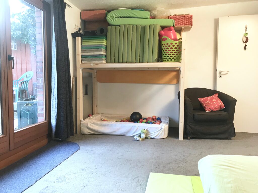 Links ist das große Fenster und die Tür zum Garten zu erkennen. Es hängt ein grau-gemusterter Vorhang an der Seite. Mittig des Bildes ist ein großes Holzregal mit einer großen Ebene für die Matratzen und Turn- und Spielmaterial zu sehen. Darunter ist Platz für ein Bällebad mit weicher Schaumstoffumrandung. Rechts neben dem Regal steht ein kleiner schwarzer Sessel der ein wenig vor einer offenstehenden Tür steht.
