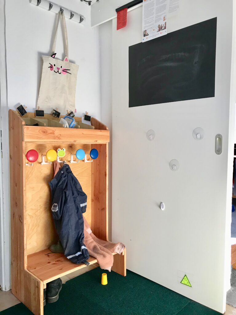 Hier ist eine kleine, liebevoll selbstgebaute Kindergarderobe aus Holz zu sehen. Die Haken sind in fünf verschiedenen Farben angestrichen und in Kinderhöhe angebracht, so dass die Kinder Ihre Kleidung selbst aufhängen können. Den oberen Abschluss der Garderobe bilden fünf kleine Fächer, in die z.B. Mützen, Brotdosen und anderes von den Kindern verstaut werden kann. Außerdem ist noch die Schiebetür zu erkennen, die den Schlaf- und Wickelraum abtrennt.